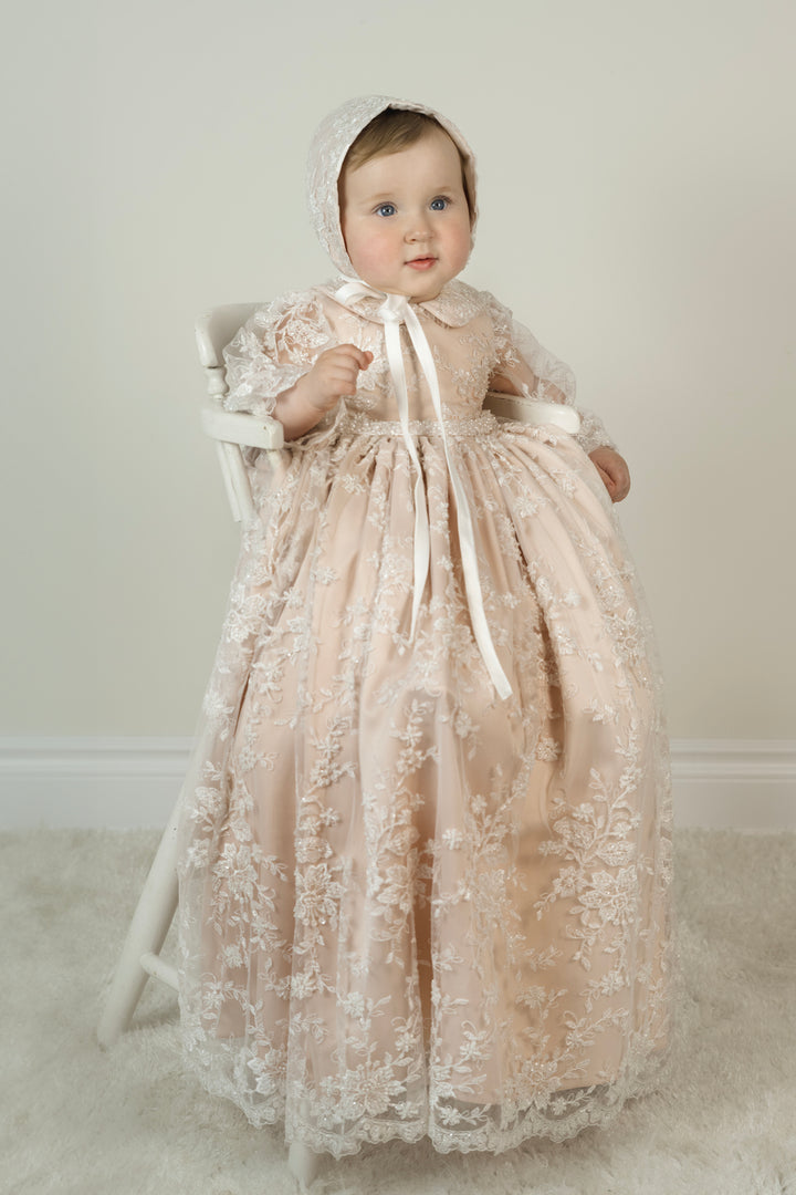 Baby girl in blush Azalea Christening Gown – long sleeve lace baptism dress with scalloped hem and matching bonnet.