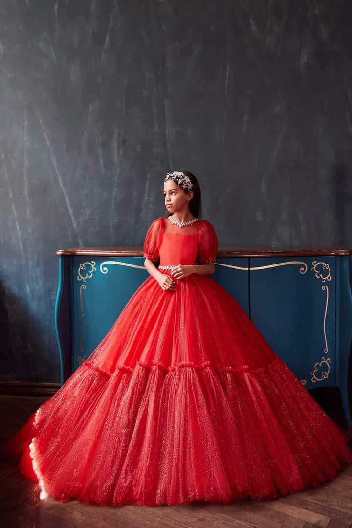 Girl wearing an elegant red Christmas party dress with puff sleeves and sparkling tulle skirt, standing in front of a vintage blue dresser by Mia Bambina Boutique