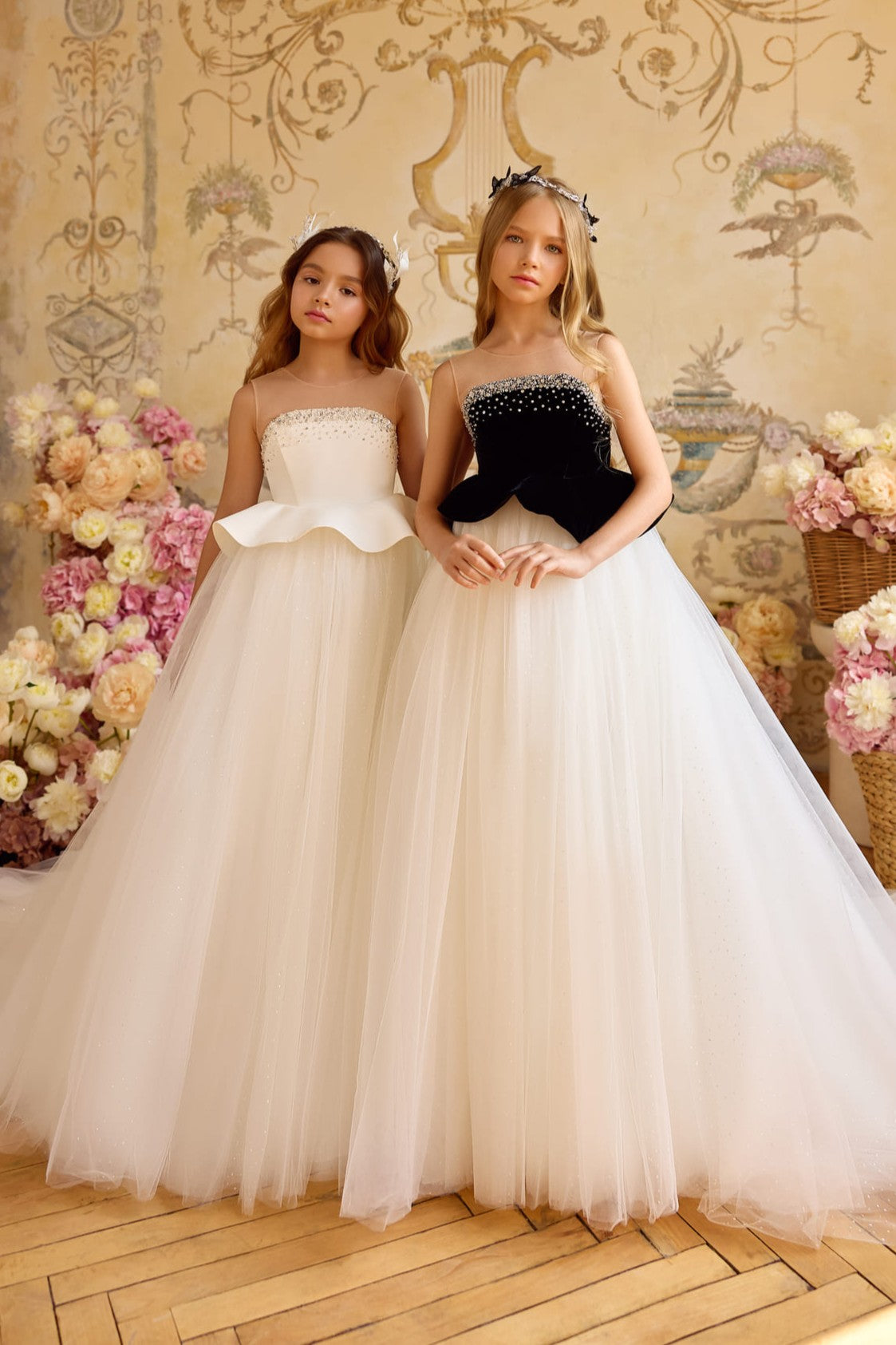 Two girls wearing elegant full-length ball gowns with sparkling tulle skirts—one with a black velvet bodice and the other in ivory satin—standing among pastel floral arrangements.
