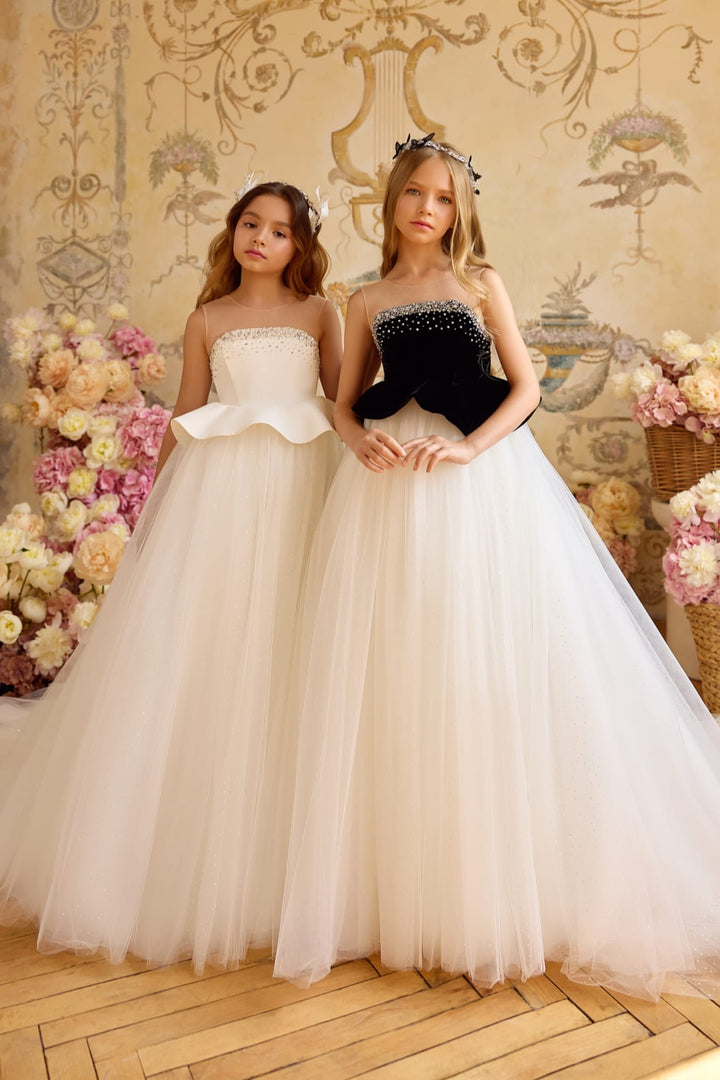Two girls wearing elegant full-length ball gowns with sparkling tulle skirts—one with a black velvet bodice and the other in ivory satin—standing among pastel floral arrangements.