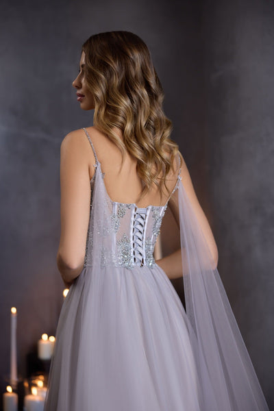 A model wearing a floor-length silver grey corset gown with a sequin bodice, illusion insert, and flowing tulle skirt, styled for prom or formal evening wear.