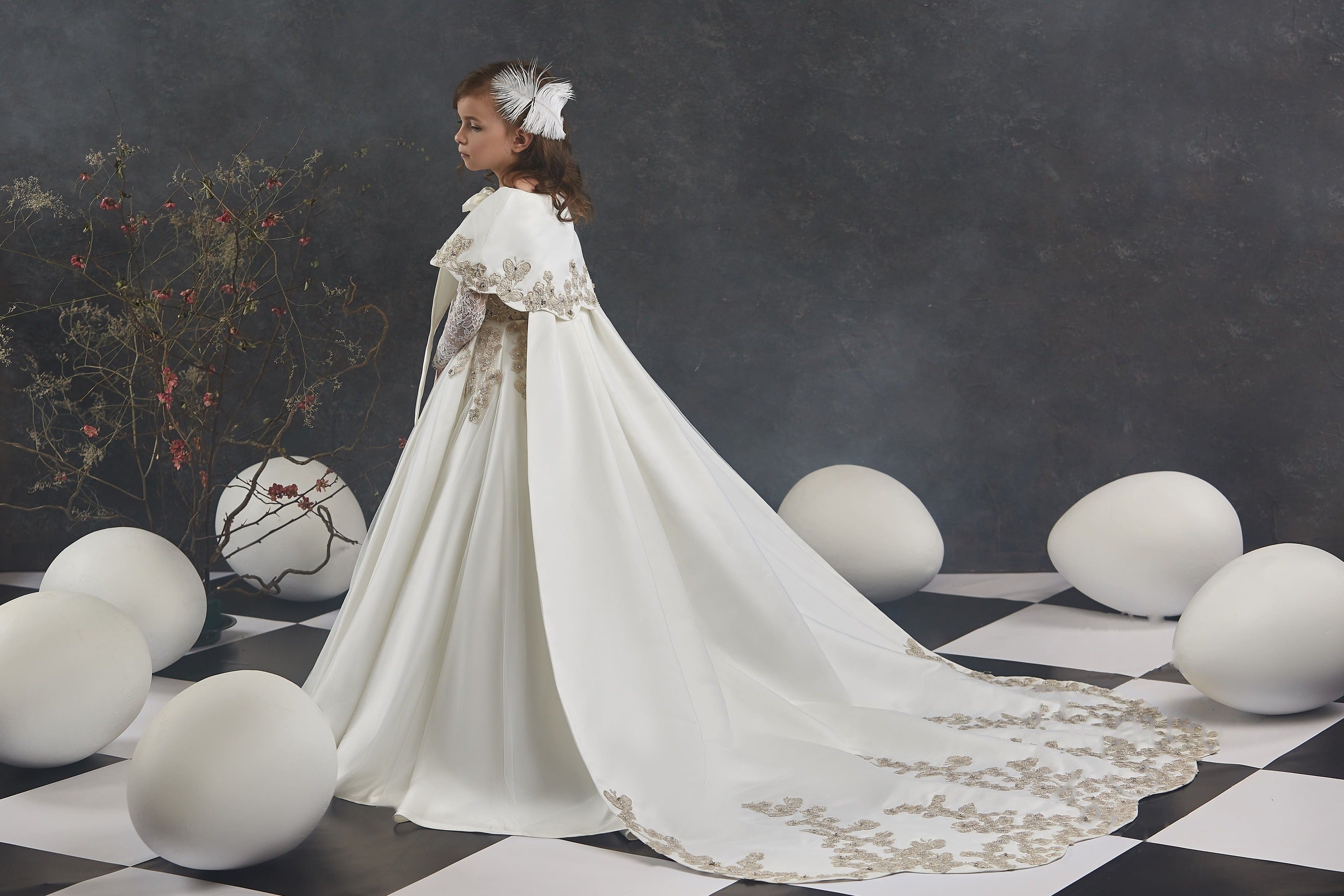 Girl wearing an ivory satin formal cape with gold embroidery, styled over a gown with long train.