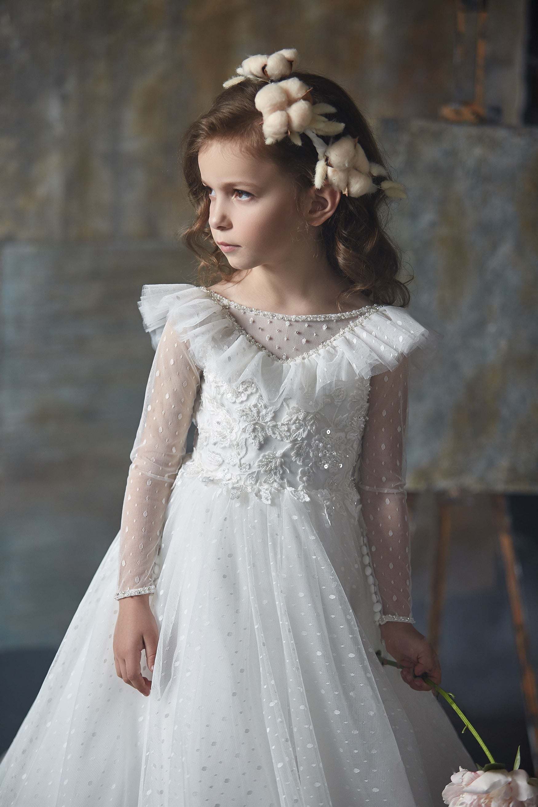 Young girl in Girls Ruffled Shoulder Open Back Tulle Dress with floral headband against a textured wall.