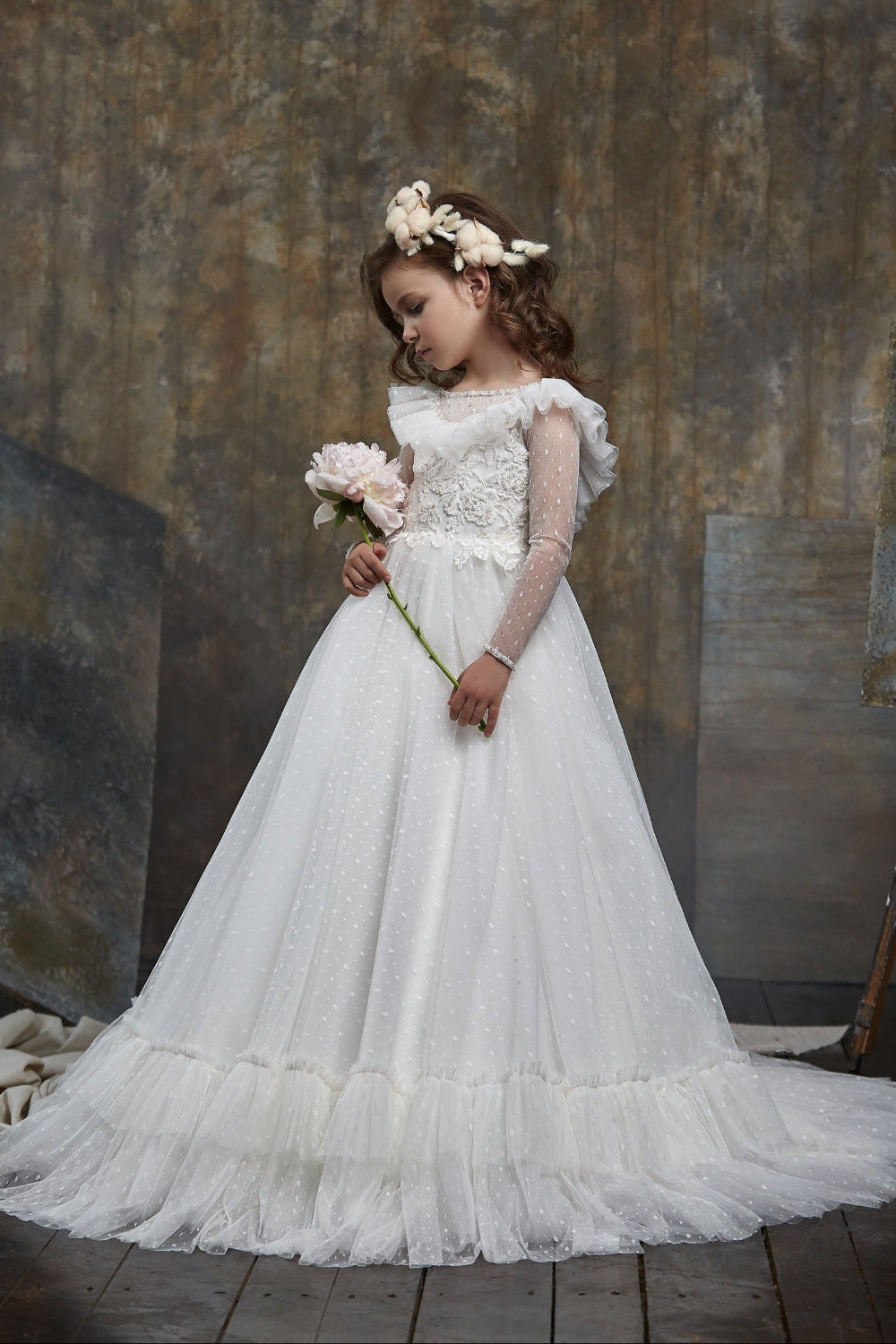 Young girl in Girls Ruffled Shoulder Open Back Tulle Dress holding flowers against a textured wall.