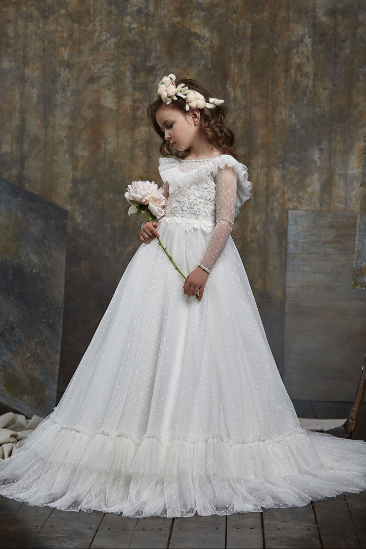 Young girl in Girls Ruffled Shoulder Open Back Tulle Dress holding flowers against a textured wall.