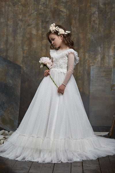 Young girl in Girls Ruffled Shoulder Open Back Tulle Dress holding flowers against a textured wall.