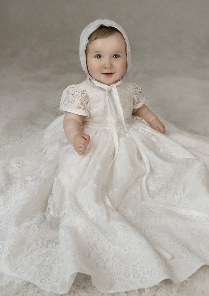 Baby girl wearing Iris Christening Gown – short-sleeved lace baptism dress with scalloped hem and matching lace bonnet.