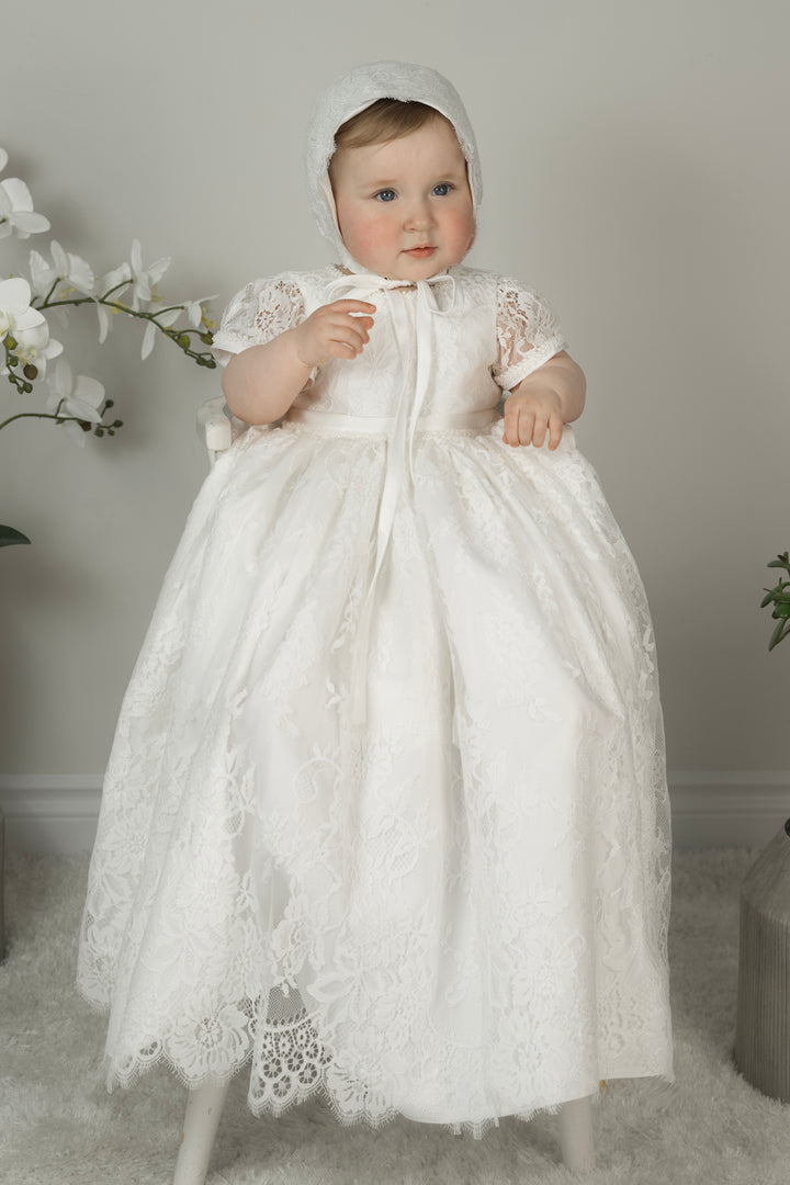 Baby girl wearing Iris Christening Gown – short-sleeved lace baptism dress with scalloped hem and matching lace bonnet.