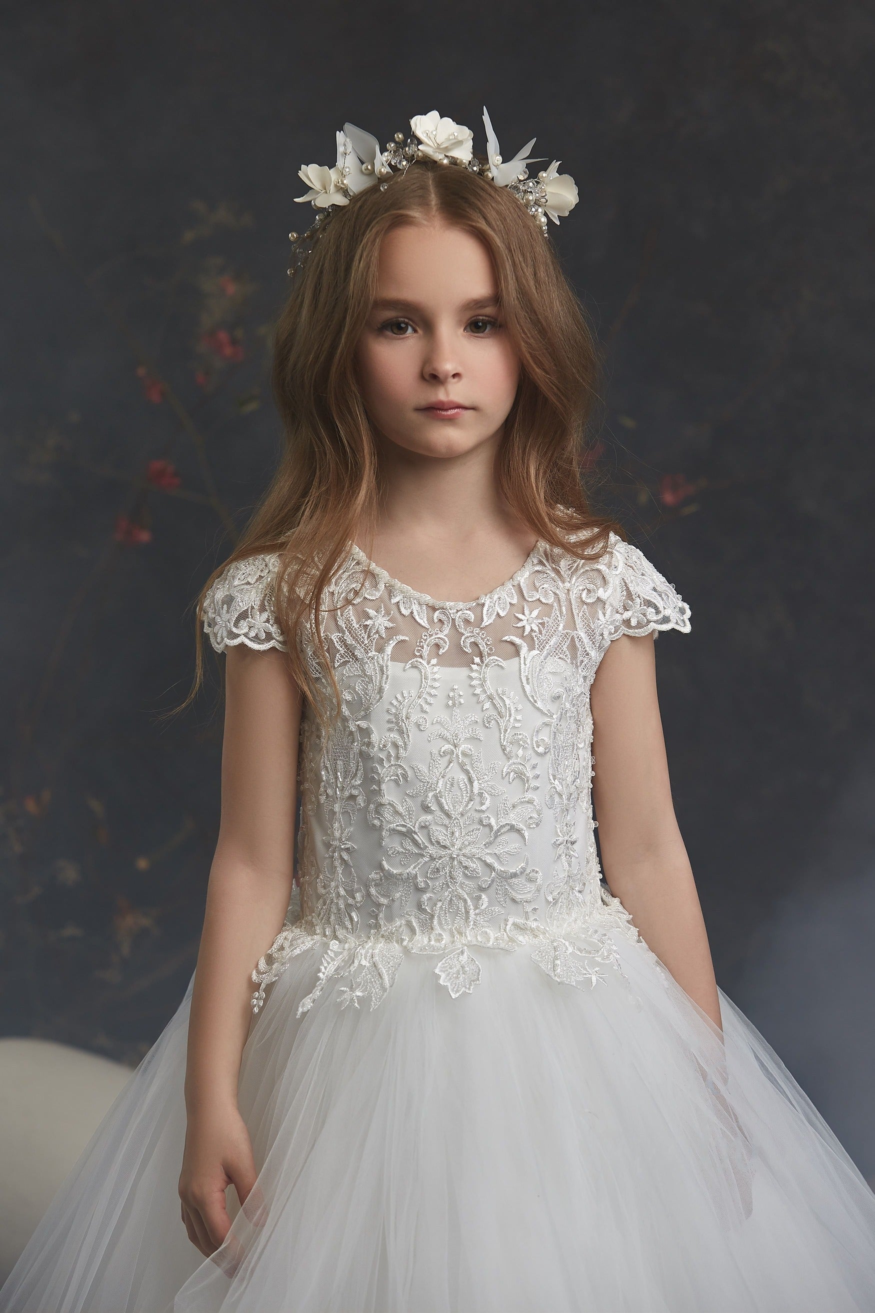 Young girl in Lace and Pearls Ivory Flowergirl Dress with floral headpiece against a dark background