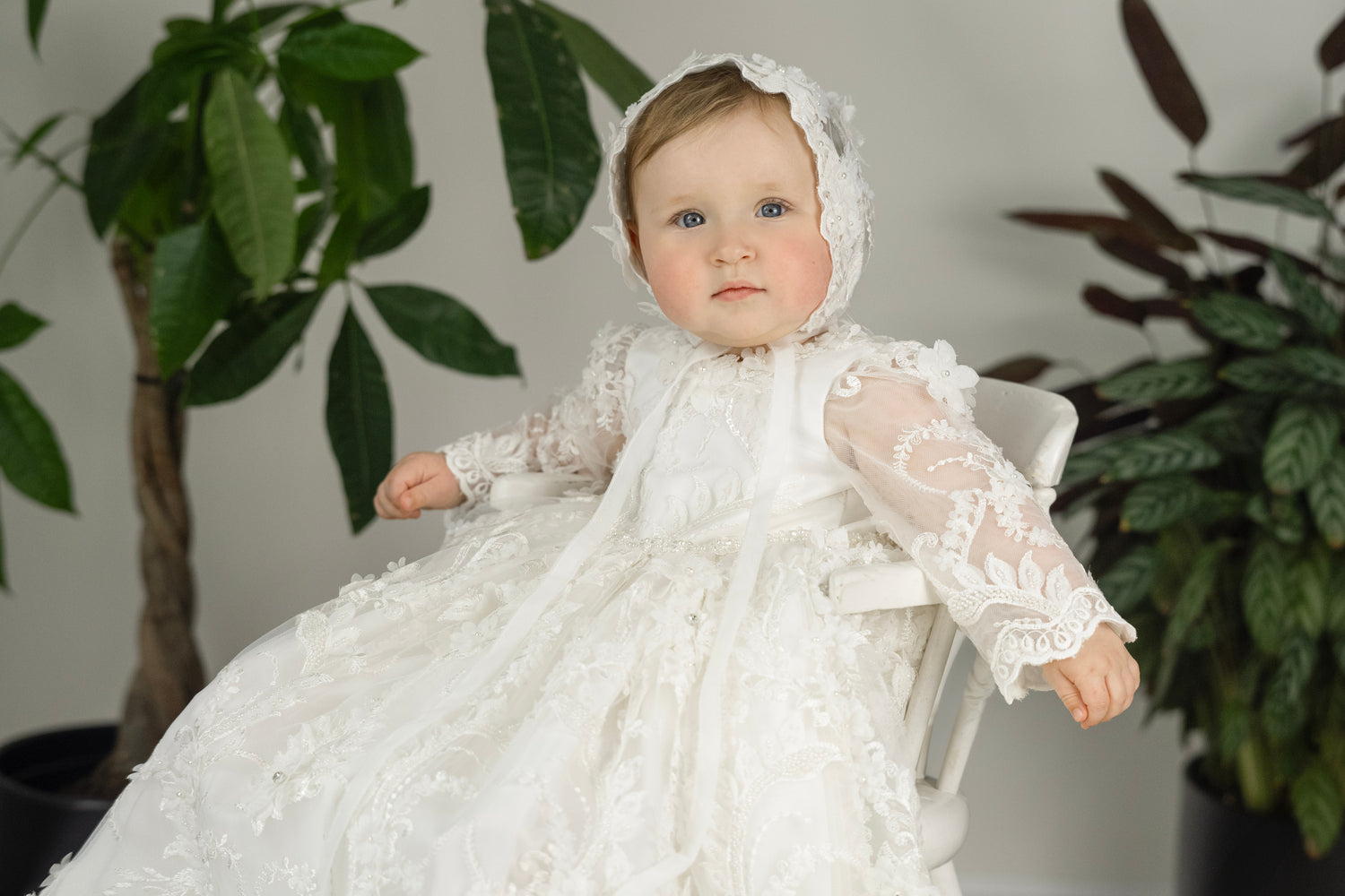 Baby girl in Lily Christening Gown – long sleeve baptism dress with floral lace, pearls, and matching bonnet.
