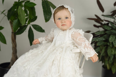Baby girl in Lily Christening Gown – long sleeve baptism dress with floral lace, pearls, and matching bonnet.