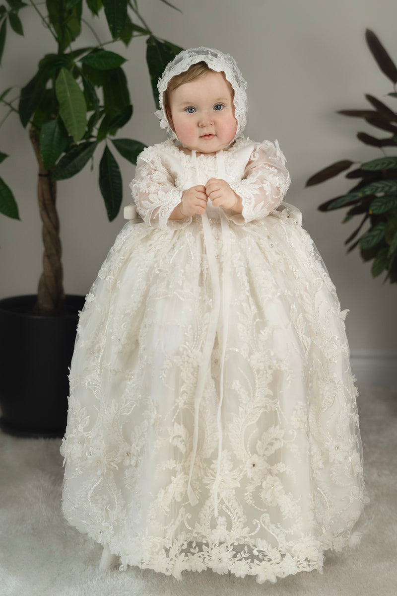 Baby girl in Lily Christening Gown – long sleeve baptism dress with floral lace, pearls, and matching bonnet.