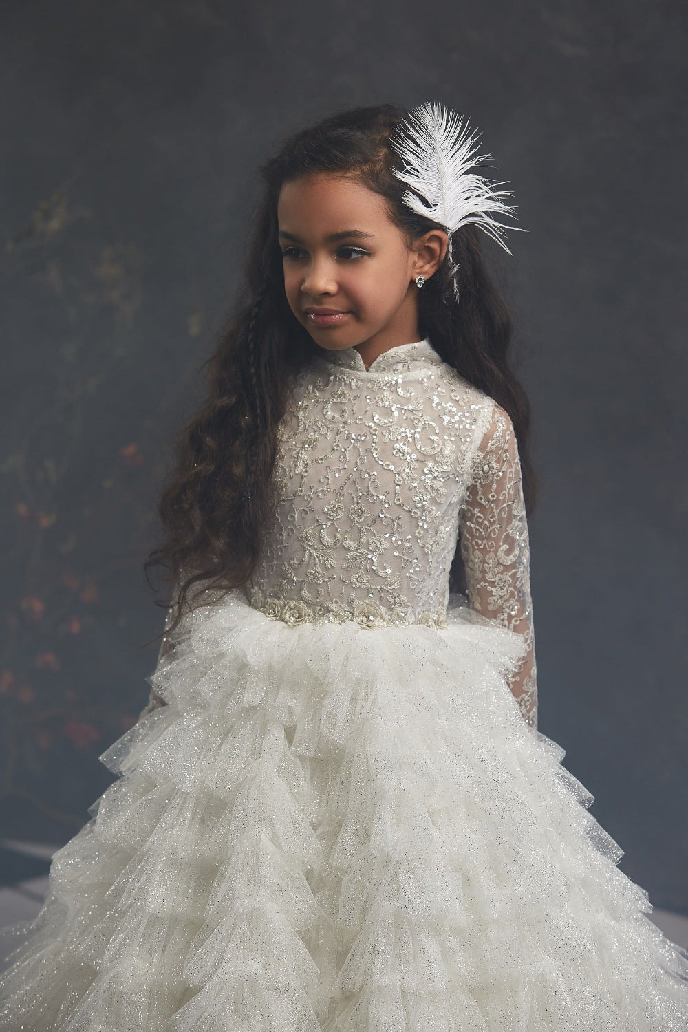 Young girl in Long Sleeve Glitter Flower Girl Dress with Ruffled skirt with a feather headpiece against a dark background