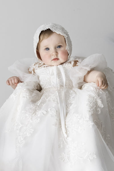 Baby girl wearing Magnolia Christening Gown with embroidered lace details, tulle skirt, puffed sleeves, and matching lace bonnet.