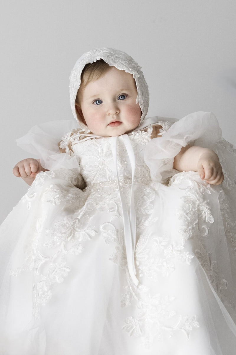 Baby girl wearing Magnolia Christening Gown with embroidered lace details, tulle skirt, puffed sleeves, and matching lace bonnet.