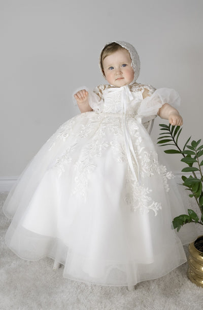 Baby girl wearing Magnolia Christening Gown with embroidered lace details, tulle skirt, puffed sleeves, and matching lace bonnet.