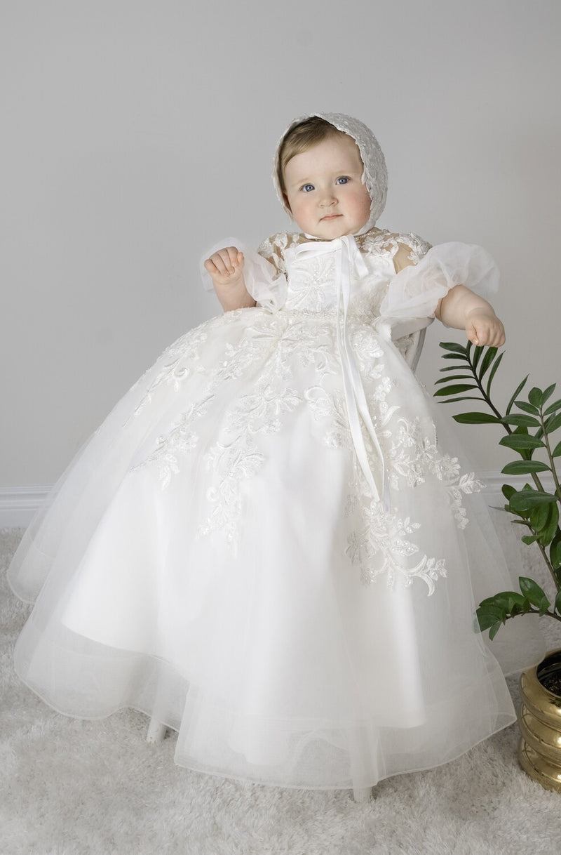 Baby girl wearing Magnolia Christening Gown with embroidered lace details, tulle skirt, puffed sleeves, and matching lace bonnet.