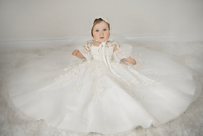 Baby girl wearing Magnolia Christening Gown with embroidered lace details, tulle skirt, puffed sleeves, and matching lace bonnet.