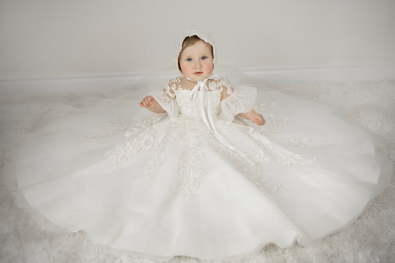Baby girl wearing Magnolia Christening Gown with embroidered lace details, tulle skirt, puffed sleeves, and matching lace bonnet.
