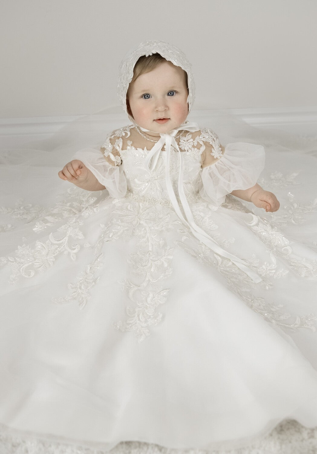 Baby girl wearing Magnolia Christening Gown with embroidered lace details, tulle skirt, puffed sleeves, and matching lace bonnet.