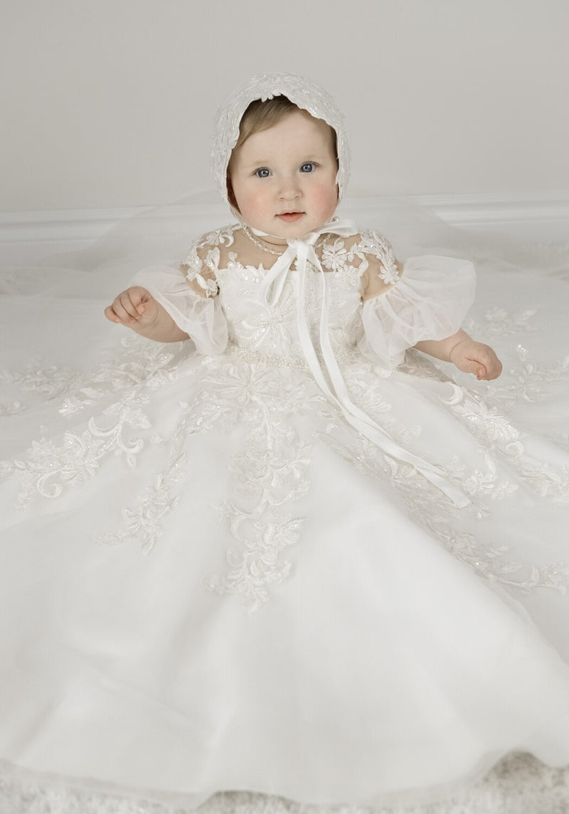 Baby girl wearing Magnolia Christening Gown with embroidered lace details, tulle skirt, puffed sleeves, and matching lace bonnet.