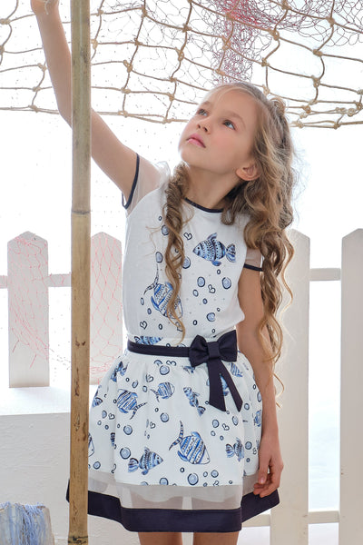 Young girl wearing a white dress with blue fish pattern, standing near a white picket fence.