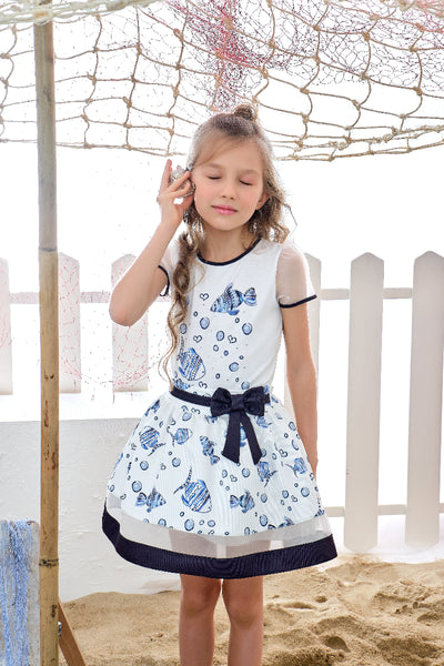 Young girl wearing a white dress with blue patterns and a navy belt, standing on sand with a white picket fence in the background.