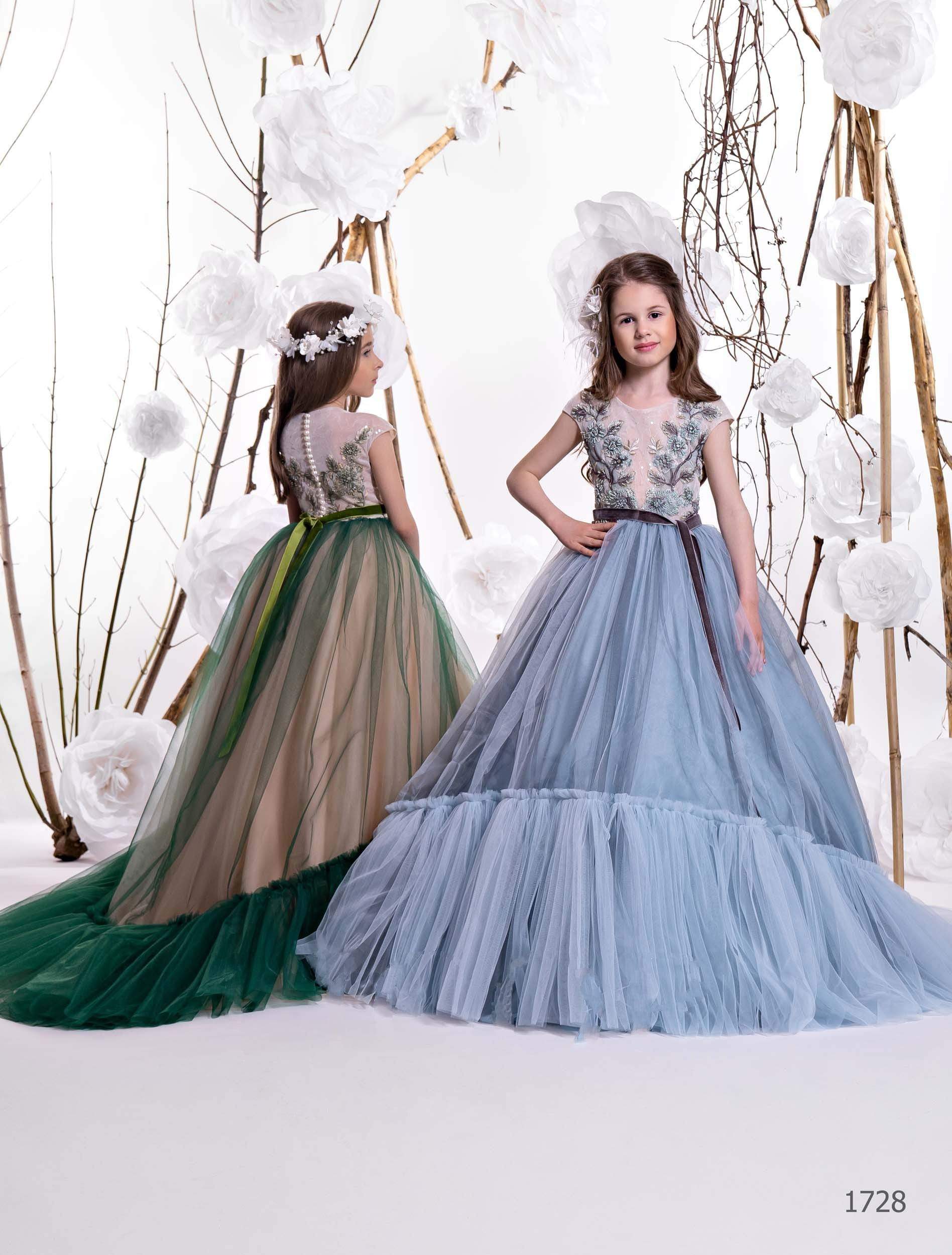 Two girls in embroidered pageant gowns – one in moss green, one in light blue – with full tulle skirts and floral bodices.